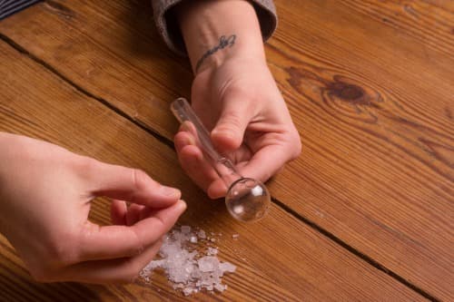 two female hands with one hand holding a crack pipe and the other hand is above crack rocks over a wooden table